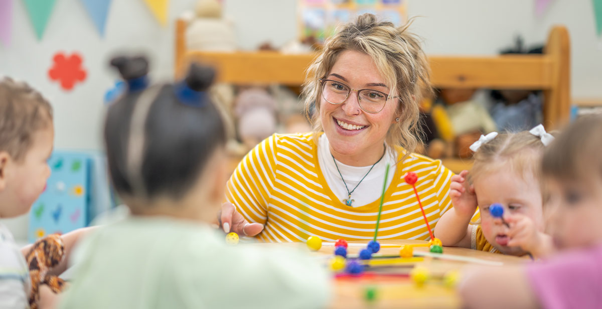 A woman in glasses and a yellow shirt sits at a table engaging with children.